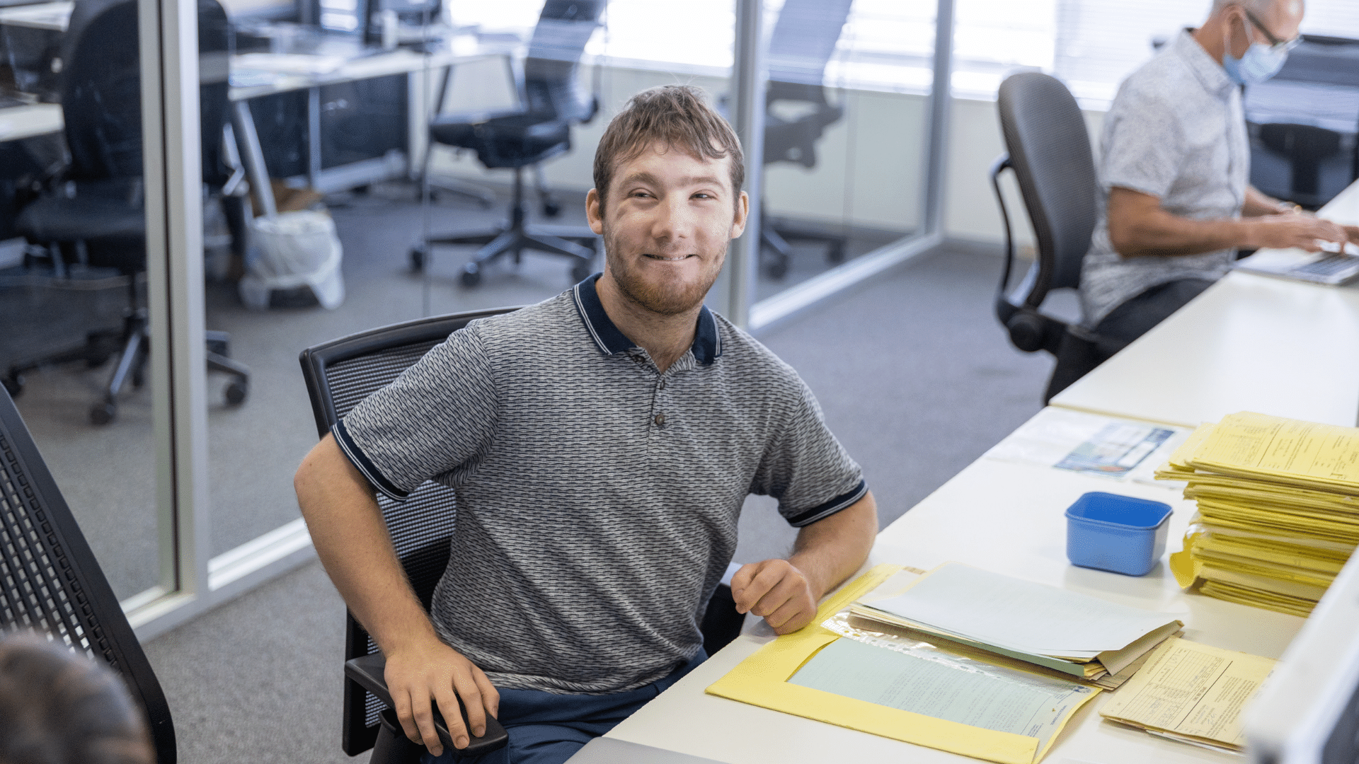 Jigsaw trainee sits at desks with papers in front of him. He's smiling at the camera.