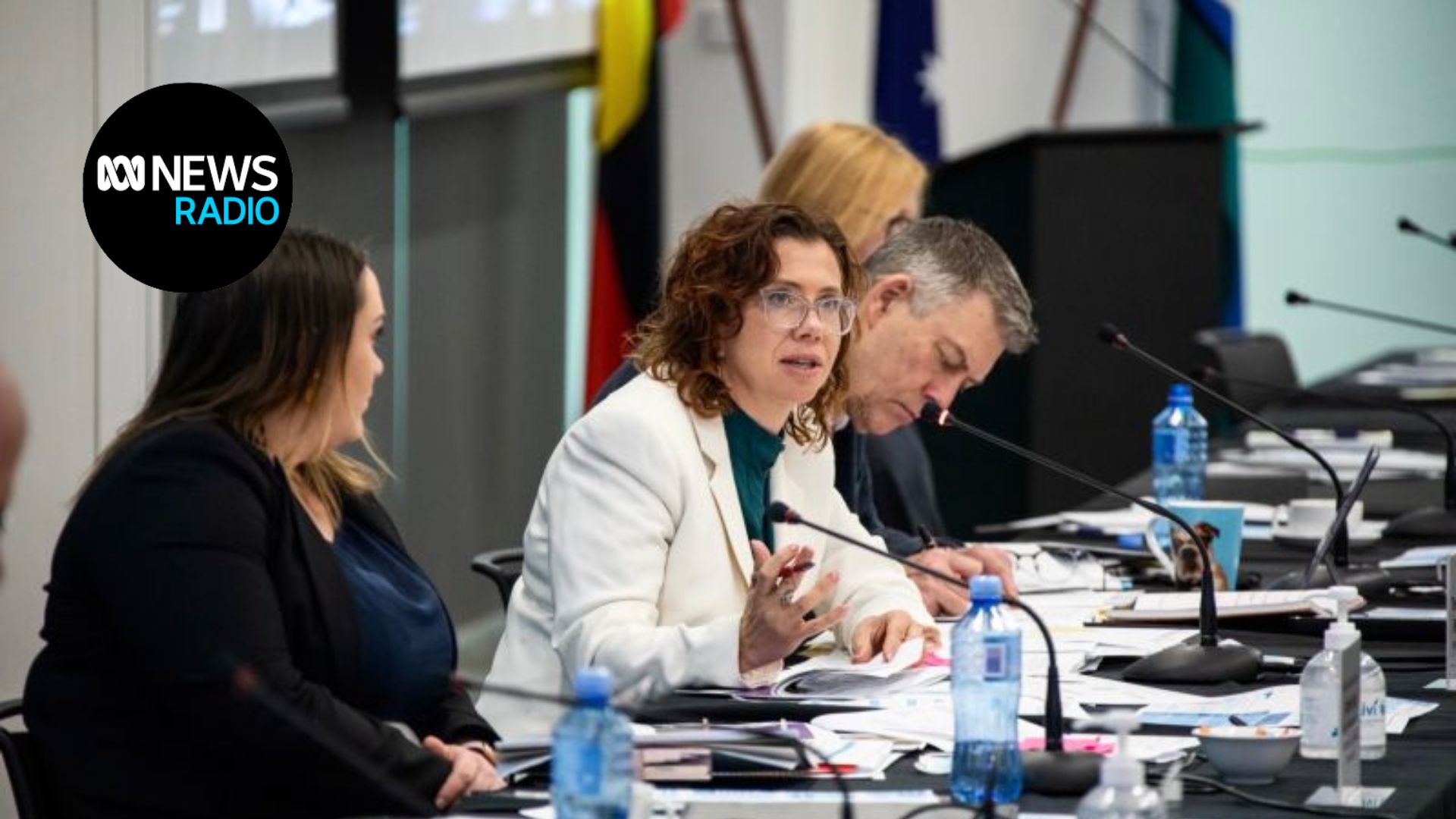 Minister Amanda Rishworth is sitting at a desk at a conference. She is wearing a cream blazer. There is a woman and man sat either side of her. The desk is covered in papers and microphones.