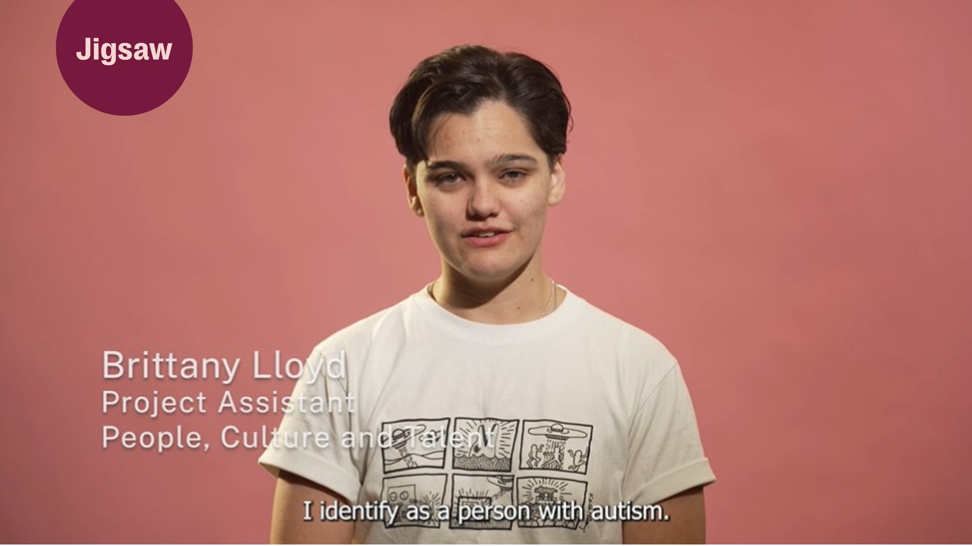 Britt Llloyd stands in front of a coral coloured background. She has short dark hair and is wearing a white t-shirt.