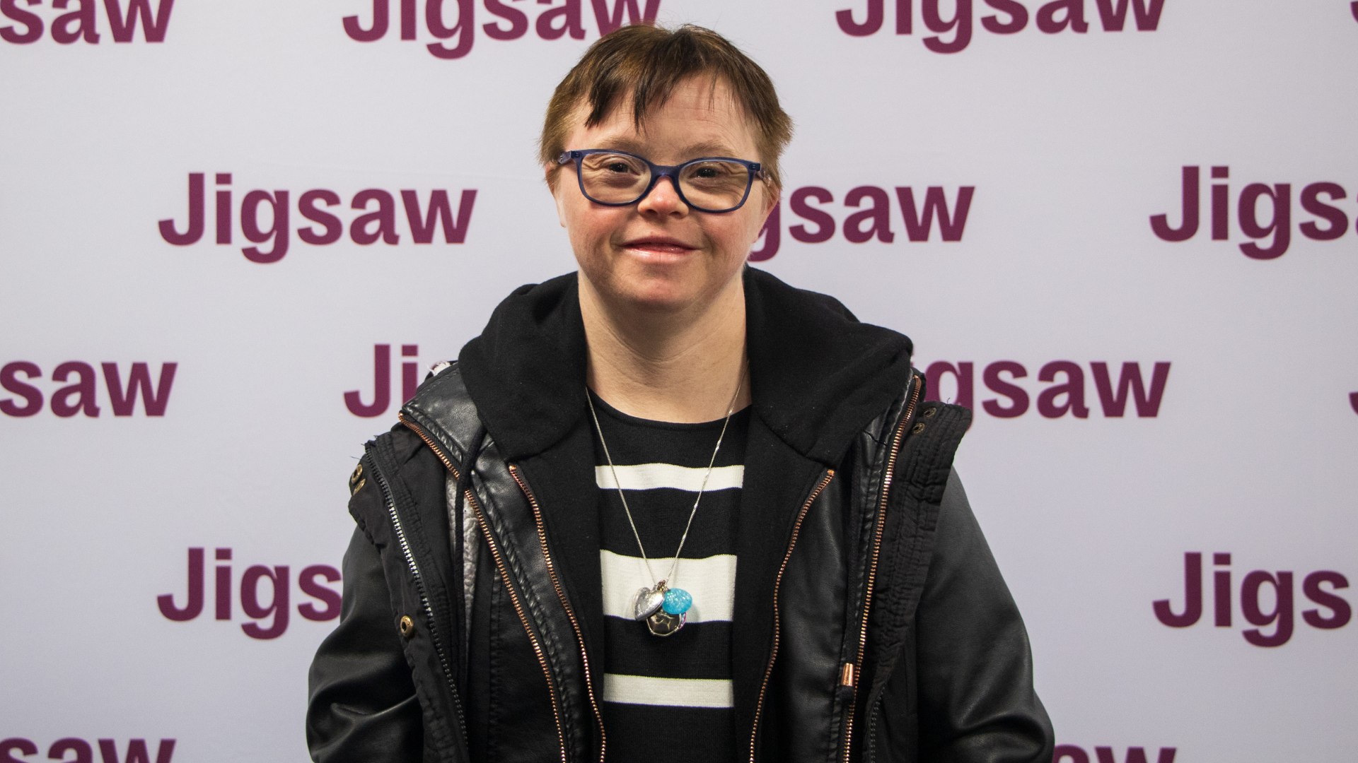 Therese smiles at camera. She has short brown hair and wears glasses. THere is a Jigsaw logo repeated behind her on a screen.